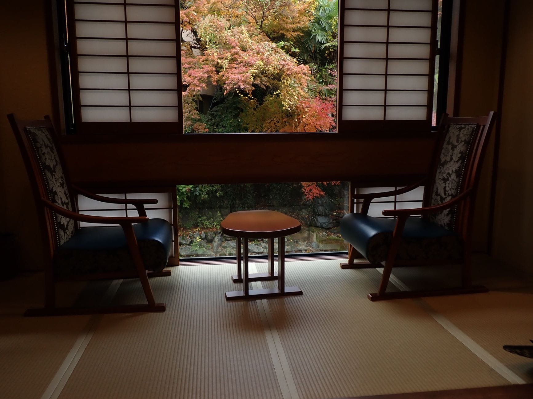 A tatami guest room with chairs by a window overlooking autumn leaves